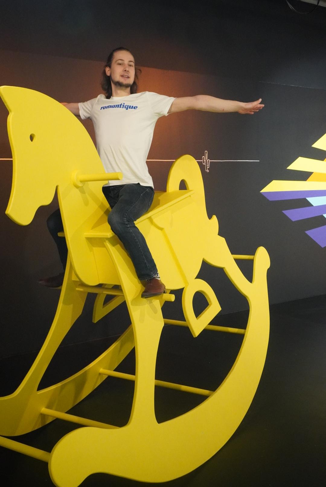 Im Museum of Popcorn in Hamburg Hafencity steht dieses gelbe übergroße Schaukelpferd. Auf dem Foto sieht man einen jungen Mann in weißen T-Shirt und Jeans auf dem Schaukelpferd freihändig balancieren. Der Hintergrund ist größtenteils schwarz.