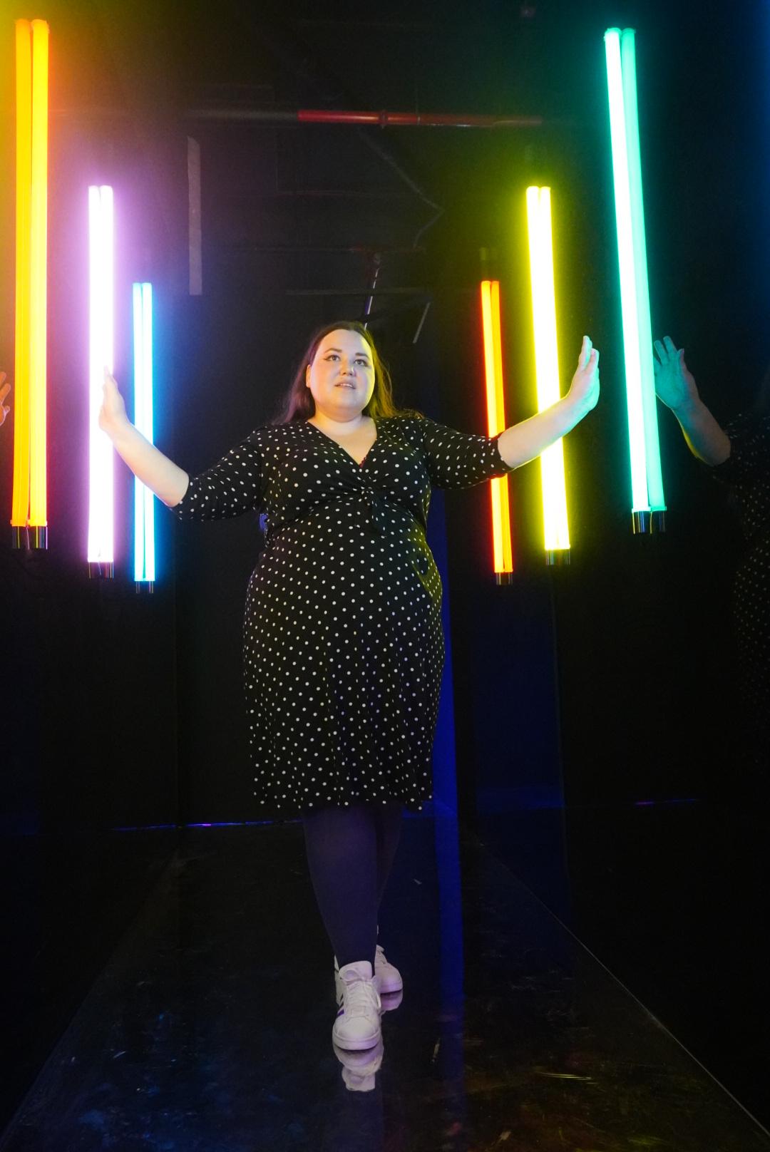 Auf dem Bild ist eine Frau in einem schwarzen Kleid mit Punkten. An der Seite des Ganges sind beidseitig bunte Lichtstangen an der Wand festgemacht.