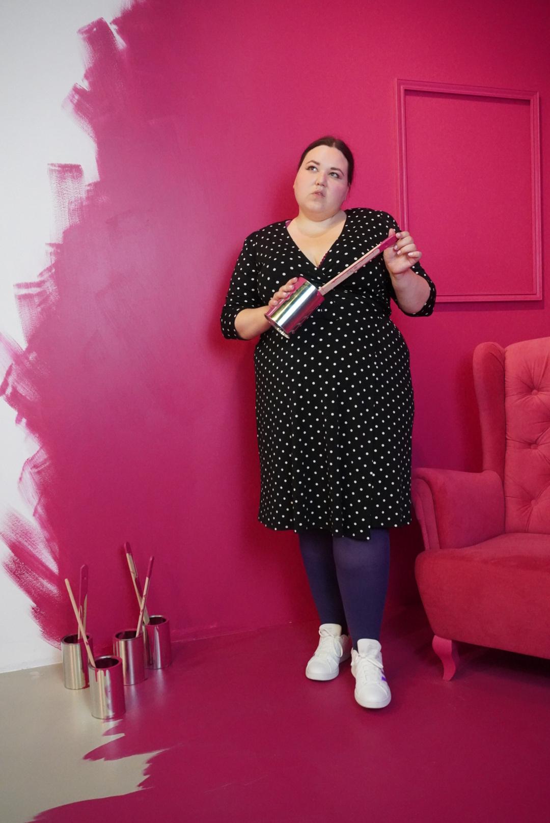 Auf dem Bild sieht man eine Frau in einem Punktkleid mit lila Strumpfhose und weißen Schuhen, die in einem komplett pinkfarbenen Raum steht, der an der linken Seite noch nicht fertig gestrichen ist. Unten auf dem Boden auf der linken Seite stehen einige silberne Farbdosen mit Pinseln drin. Die Frau hält eine der Dosen mit Pinsel in den Händen.