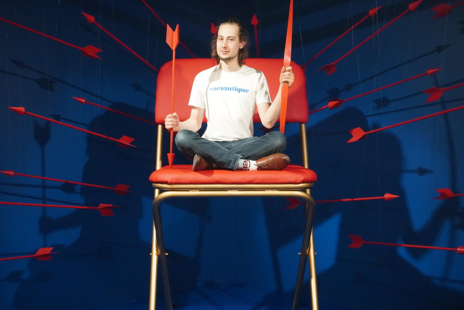 Auf dem Bild sieht man einen jungen Mann in weißen T-Shirt und Jeans in einer Bilderkulisse im Museum of Popcorn in Hamburg Hafencity. Im Bildzentrum steht ein übergroßer Klappstuhl in Rot und Silber, der junge Mann im Schneidersitz hält in der einen Hand einen roten Pfeil aus Holz und in der anderen Hand einen roten Bogen. Um ihn herum schweben viele rote Pfeile in der Luft vor dem blauen Hintergrund.