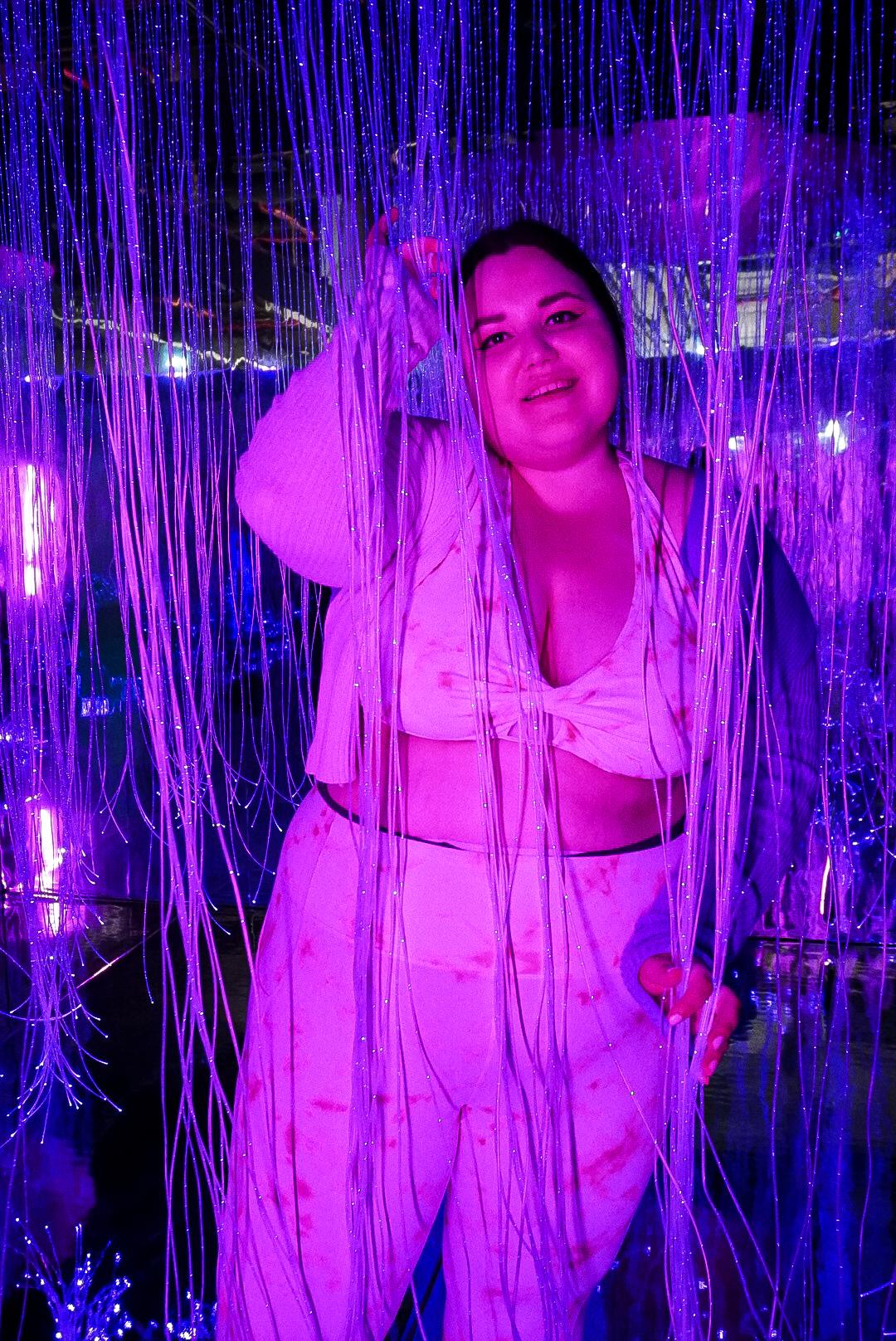 Auf dem Bild sieht man eine junge Frau im Museum of Popcorn in Hamburg Hafencity in einem Outfit aus Cadigan, Sport BH und Sporthose. Die Frau und der gesamte Vordergrund in dem abgedunkelten Raum sind in pinkfarbenes und blaues Licht gehüllt. Die Frau hält sich mit beiden Händen an blauen Lichtfäden fest, die im ganzen Raum verteilt sind.